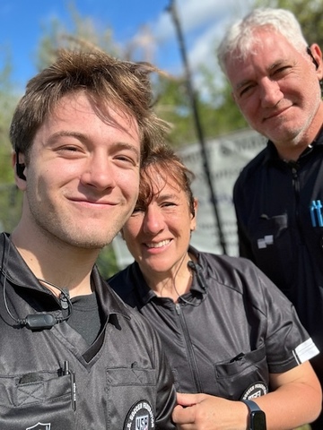 Three USSF referees in black jerseys smiling on a sunny match day